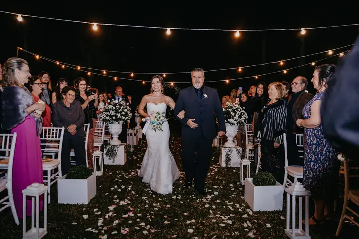 Noiva entrando com o pai sob um varal de luzes, convidados de pé