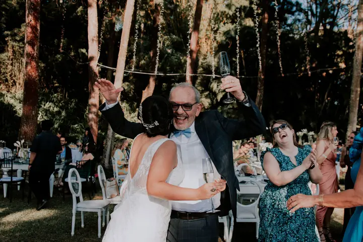 Noiva abraçando um convidado que ergue a taça na festa